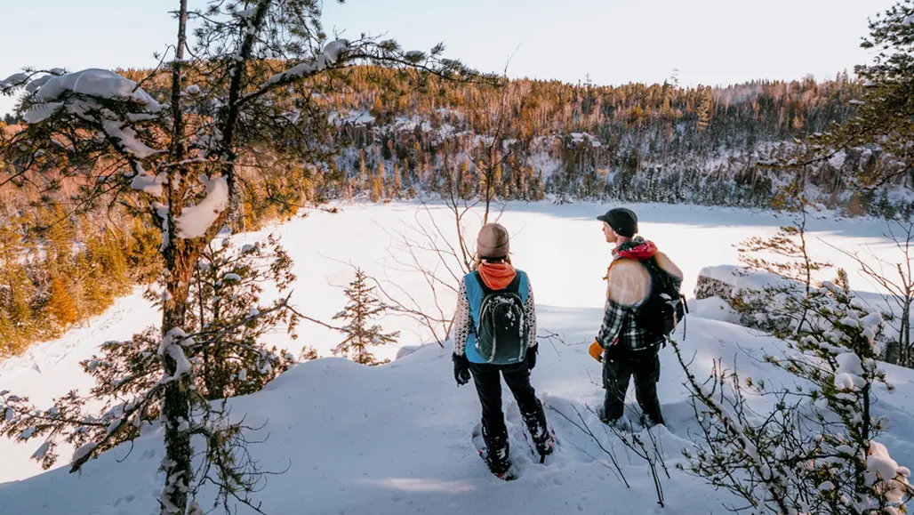 Two people snowshoeing in Northern Ontario stop to admire the view of the forest blanketed in snow across a frozen lake.