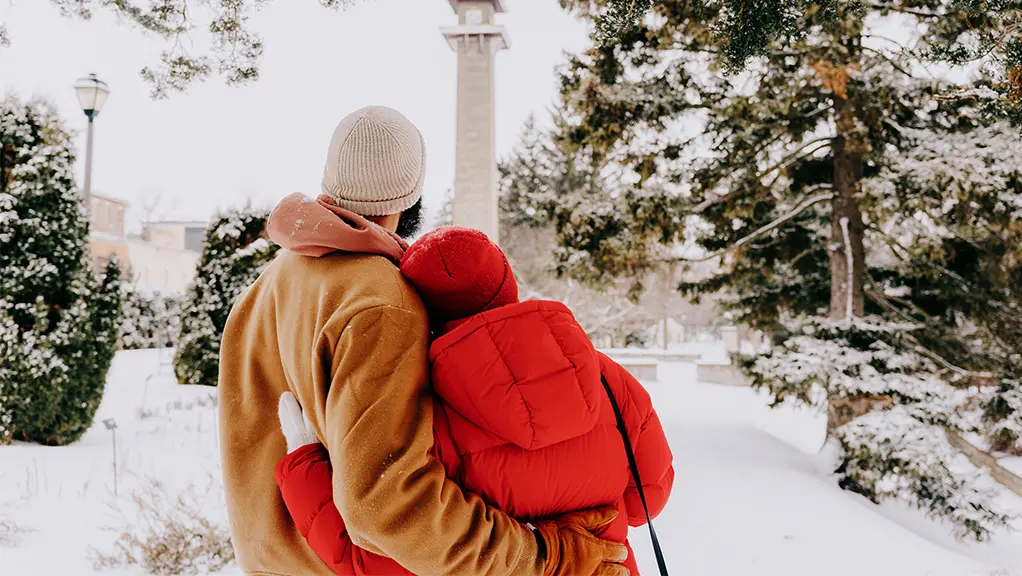 A couple embraces surrounded by a snowy landscape of trees with a tall tower off in the distance.