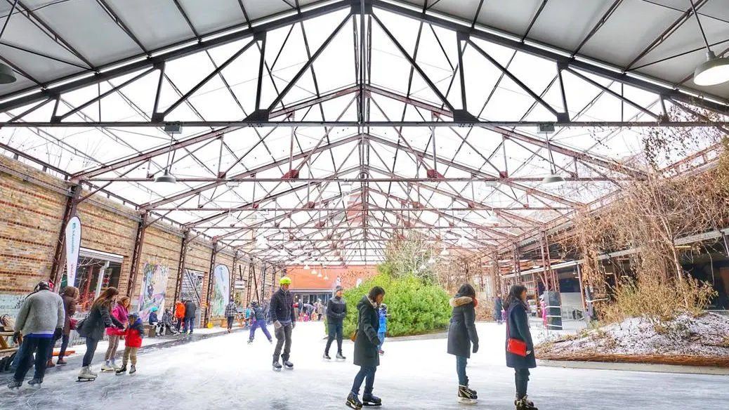 Several people enjoy ice skating on an open-air skating rink under brick factory beams at Evergreen Brickworks.