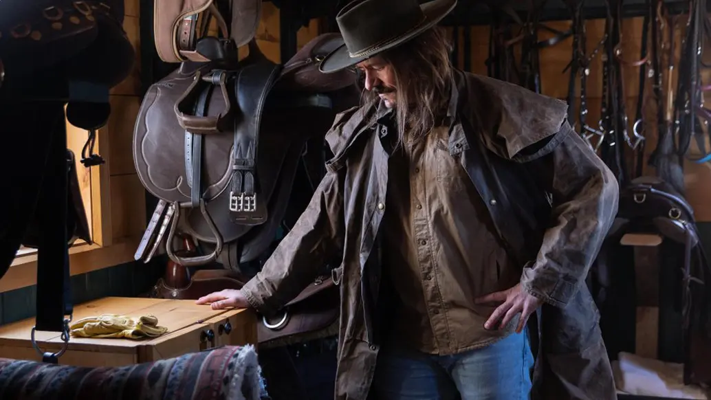 A man wearing a cowboy hat and an oilskin duster coat inspects saddles and other horseback riding gear.