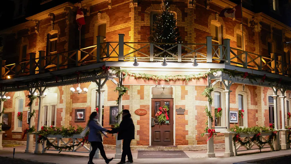 A couple holding hands stand at the front door of a boutique inn in a heritage building decorated with wreaths and bows for the holiday season.