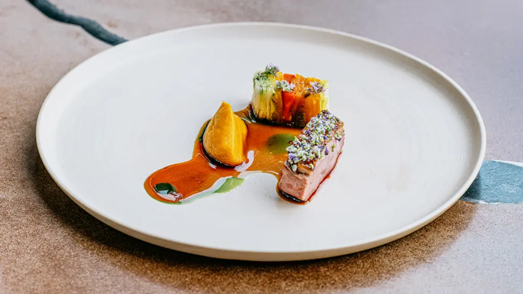 Eloquently served dish of meat and vegetables covered in sauce, with a woman's hand holding a wine glass beside it.