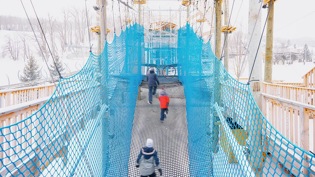 3 人の子供が、冬の風景を背景に、プラットフォームからプラットフォームへと高架ロープ コースに沿って歩いています。