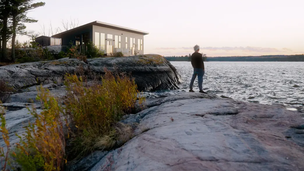 Una persona parada en una costa rocosa junto a una pequeña cabaña con vista al lago en el noroeste de Ontario.