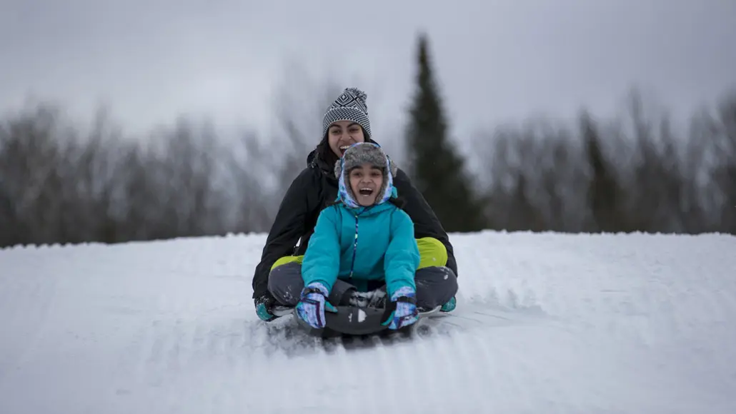 A teen and a young girl ride down a snowy hill on a toboggan at a winter resort in Muskoka.