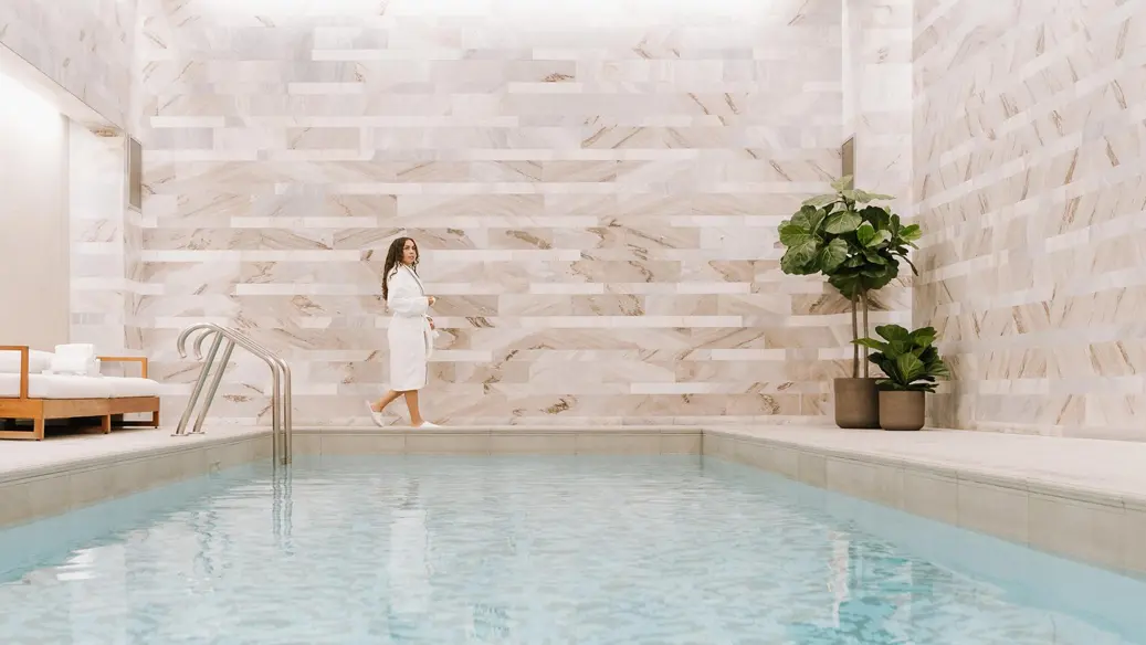 A woman in a white terry-cloth bath robe and slippers walks alongside an indoor pool at an upscale hotel spa in Toronto.
