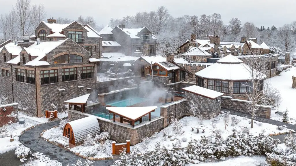 고풍스러운 석조 건물들이 가볍게 눈으로 덮인 채, 수증기가 피어오르는 온수 야외 수영장을 둘러싸고 있는 스파 리조트의 모습입니다.