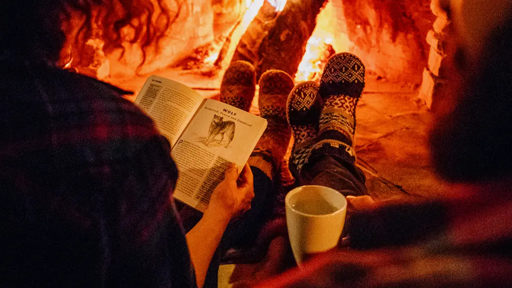 2 人の人物が、心地よい靴下を履いて、火のともった暖炉の前に並んで座ります。1 人が本を読み、もう 1 人がマグカップを持ち、暖かく親密な冬の雰囲気を醸し出しています。