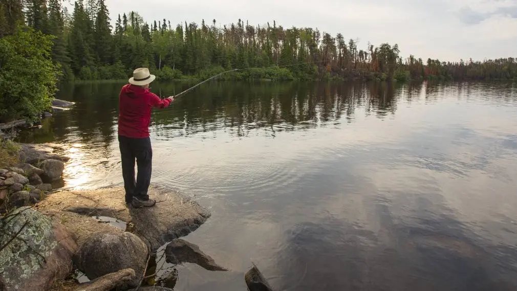 Un hombre parado en una orilla rocosa lanza un sedal a un lago tranquilo en un parque provincial de Ontario.