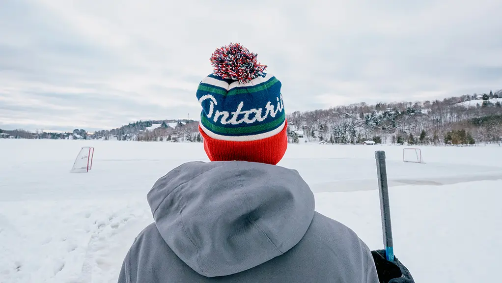 Un joven se encuentra frente a una pista de hockey sobre hielo en un estanque sosteniendo un palo de hockey.