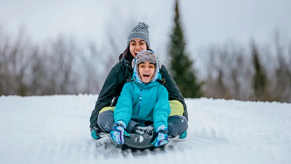 A teen and a young girl ride down a snowy hill on a toboggan at a winter resort in Muskoka.
