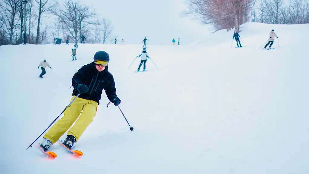 霧の日に、黄色いパンツをはいたスキーヤーが雪に覆われた斜面を滑降し、背景には他のスキーヤーが見える。