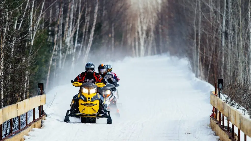 Drei Schneemobilfahrer fahren mit ihren Schlitten über eine schneebedeckte Brücke, im Hintergrund ist ein Wald zu sehen.