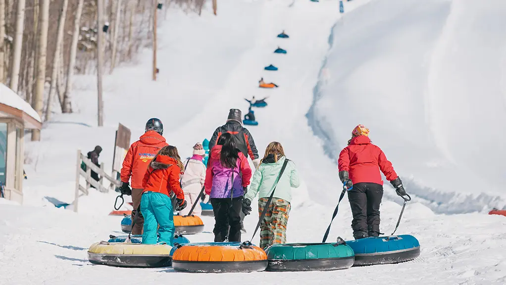 Families pull colorful snow tubes uphill at a tubing park, with snowy slopes and trees in the background.