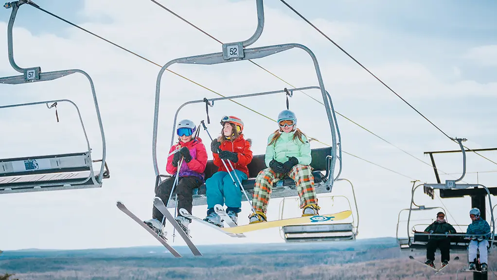冬の晴れた日に、3人がスノーボードとスキーを積んだスキーリフトに乗って雪の斜面を登っていきます。