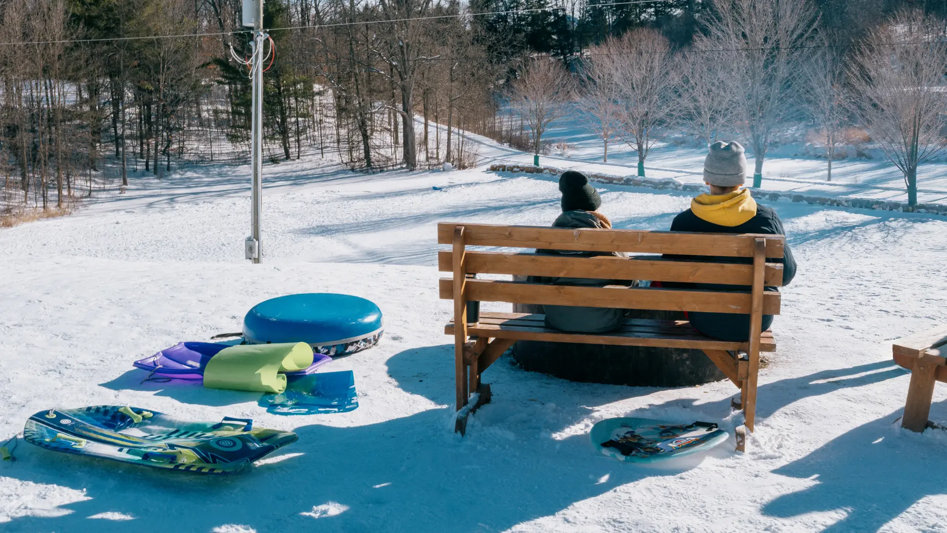 두 사람이 눈 덮인 언덕과 나무들이 내려다보이는 벤치에 앉아 있고, 그들 옆에는 썰매와 튜브가 놓여 있다.