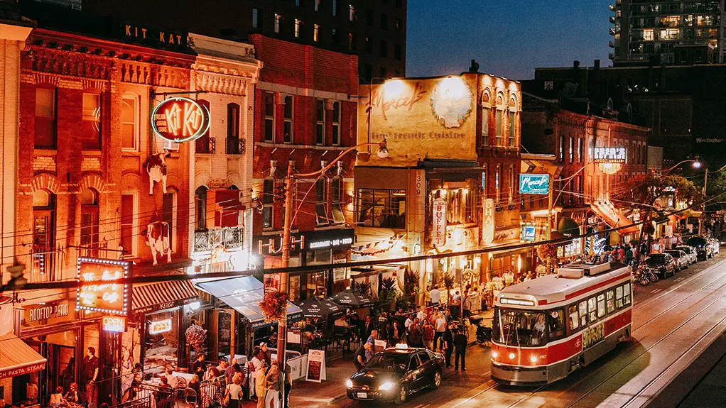 A crowded nightlife street with illuminated bars, restaurants, pedestrians and a streetcar passing through at night.
