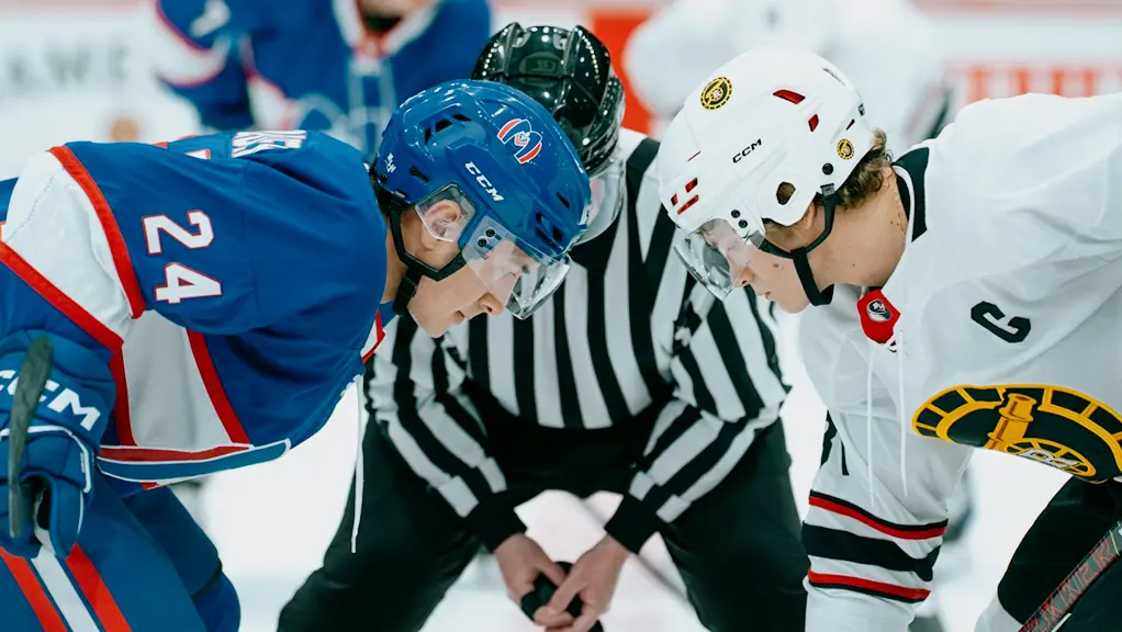 파란색 유니폼을 입은 선수와 흰색 유니폼을 입은 선수, 두 명의 하키 선수가 퍽을 차지하기 위해 마주 보고 서 있고, 심판은 그들 사이에 웅크리고 있다.