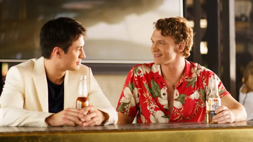 Deux hommes assis à un bar tenant des bouteilles de bière, discutant et se souriant l’un à l’autre, l’un portant un blazer blanc et l’autre, une chemise rouge à fleurs, dans un cadre moderne et chaleureux.