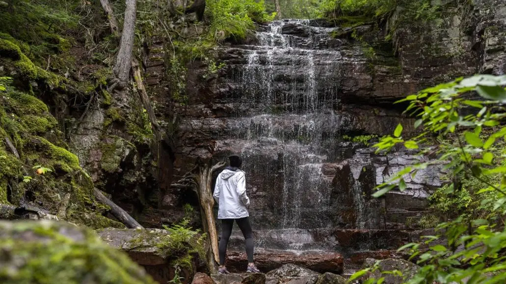Ein Wanderer hält inne, um einen sanft plätschernden Wasserfall zu bewundern, der von moosbewachsenen Felsen, Bäumen und üppigem Grün im Norden Ontarios umgeben ist.