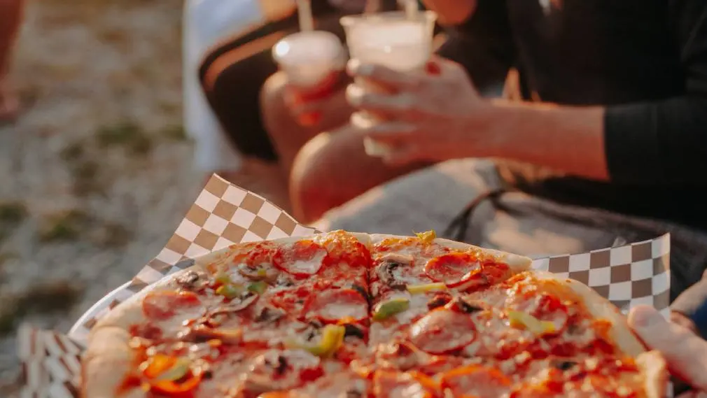 Une pizza fraîchement préparée, des gens tenant des boissons à proximité, sur la plage.