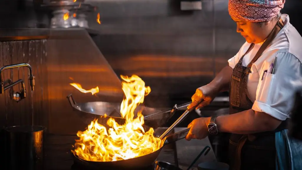 Chef cuisinière faisant habilement flamber un plat dans un grand wok, dans la cuisine du restaurant Kiin de Toronto, étoilé MICHELIN.