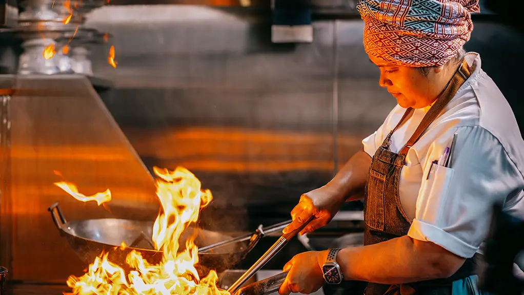 A woman chef expertly flame-cooks a dish in a large wok in the kitchen of Kiin, a MICHELIN-starred restaurant in Toronto.