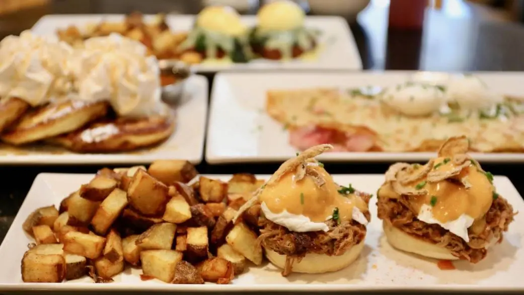 白い皿に盛られたグルメなブランチ料理の数々。プルドポークのエッグベネディクト、クリスピーポテト、ホイップクリーム添えのパンケーキなど。