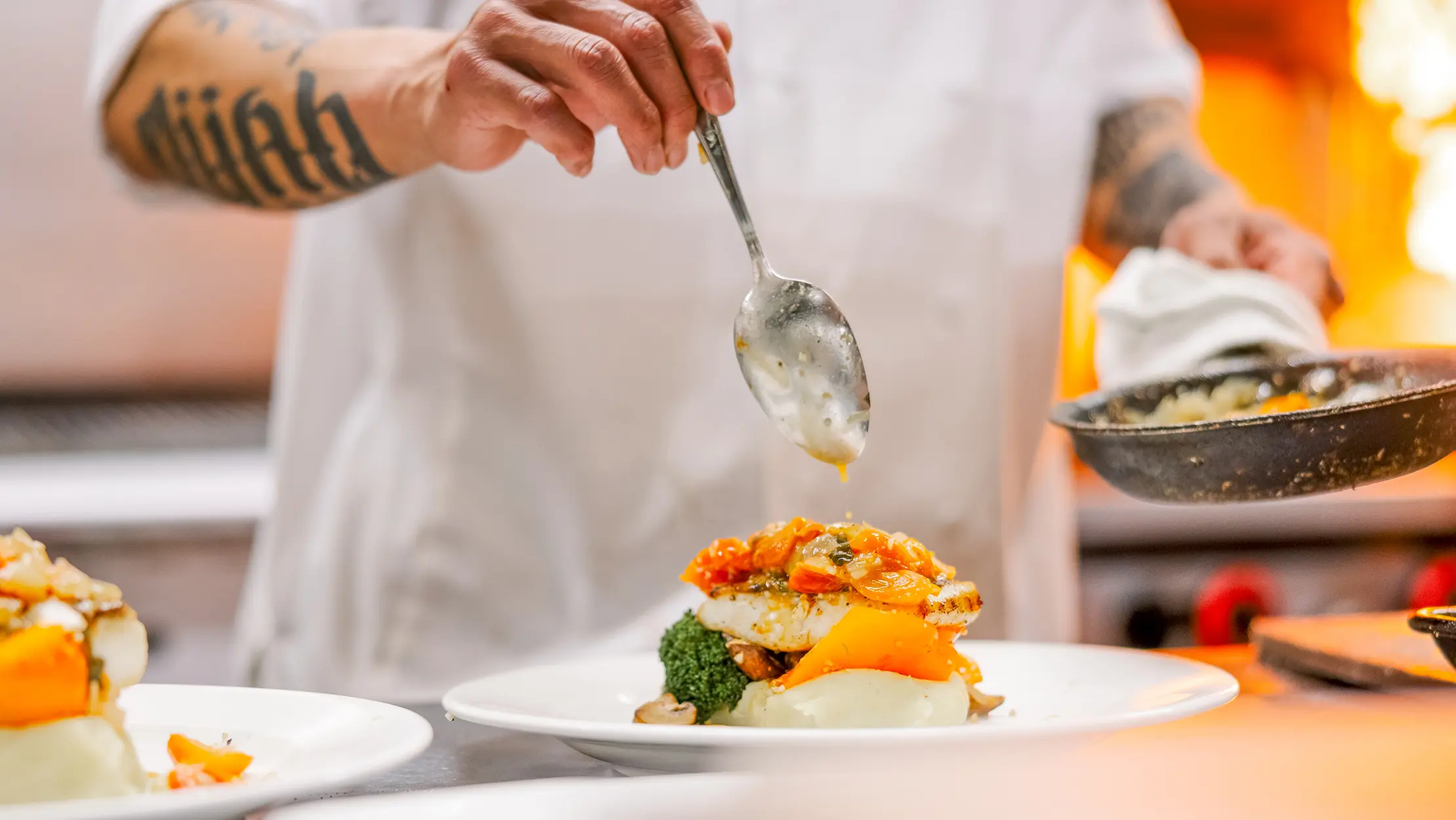 Chef plating a seafood dish with vegetables and sauce in a restaurant kitchen.