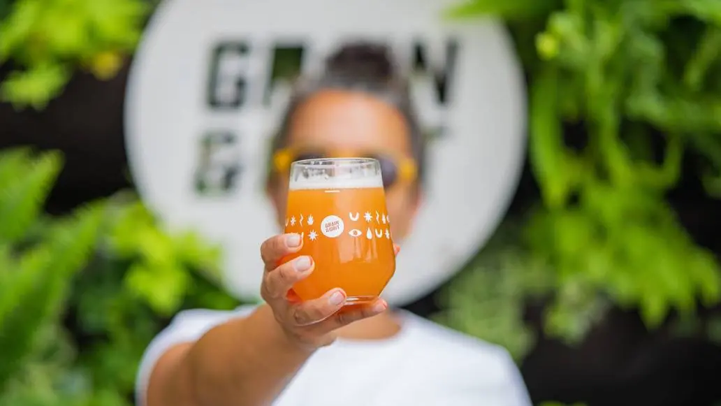 A woman holds up a freshly poured glass of craft beer, standing in front of the brewery sign.