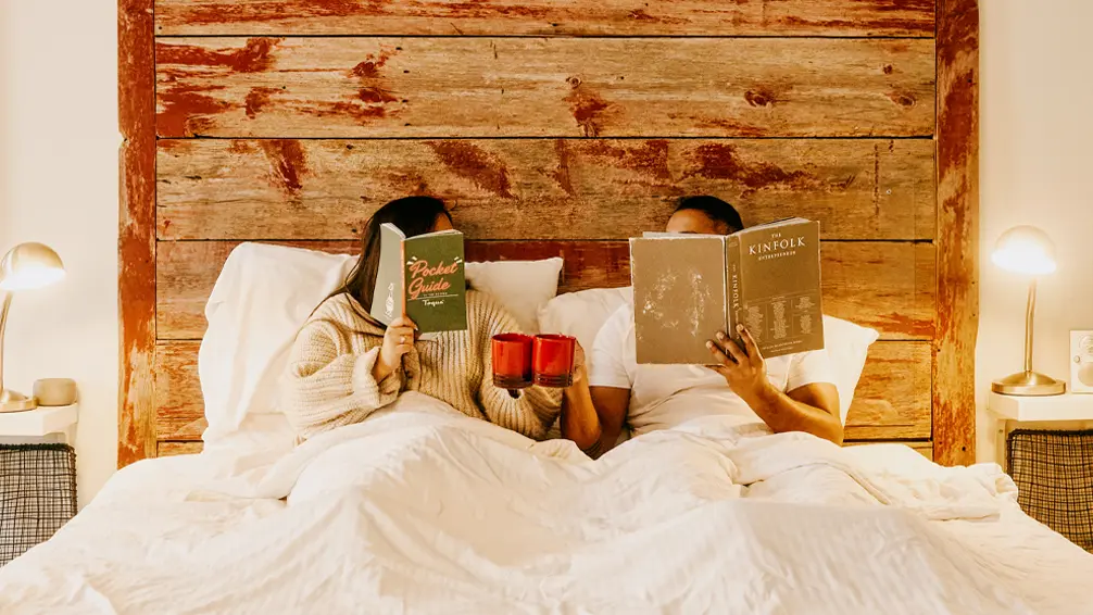 침대에 누워 책을 읽는 커플이 머그잔을 들고 있다.