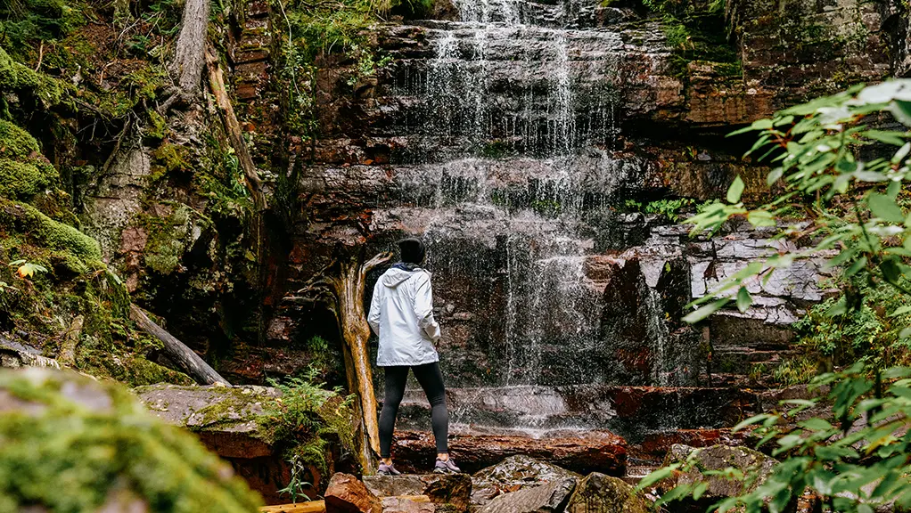A hiker stops to admire a gentle waterfall surrounded by mossy rocks, trees and lush greenery in Northern Ontario.
