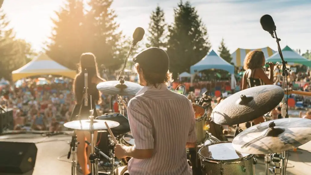 View from over the shoulder of a drummer in a band performing on stage at an open air music festival.
