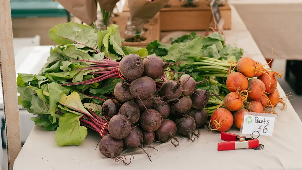 Remolachas frescas y otras verduras expuestas en una mesa en un mercado de agricultores, con flores de fondo.