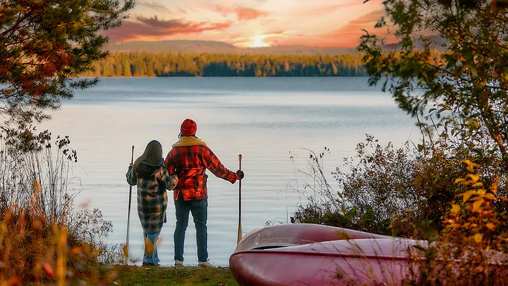 秋の紅葉に囲まれた静かな湖畔に、カヌーとパドルを持った二人が立っている。