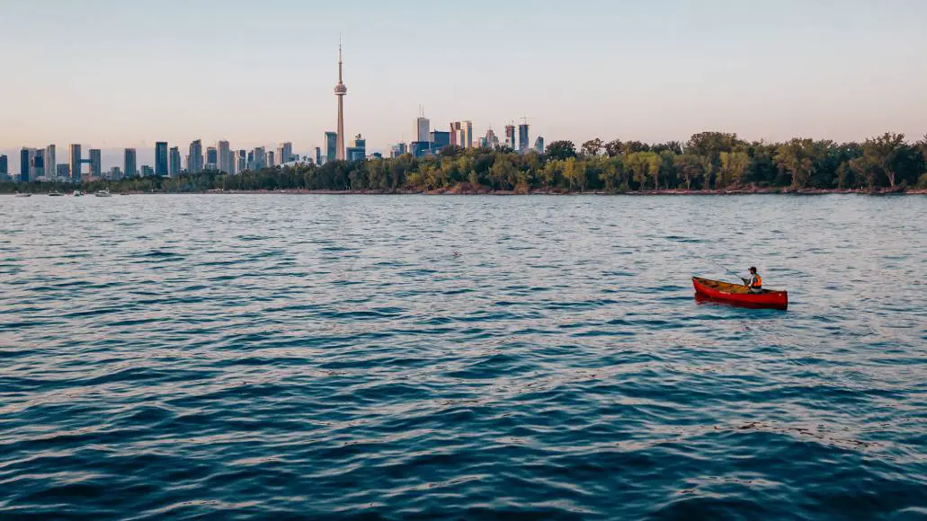 Un homme lance sa ligne de pêche depuis un canoë dans les eaux du port de Toronto, avec la Tour CN et la silhouette de la ville en arrière-plan.