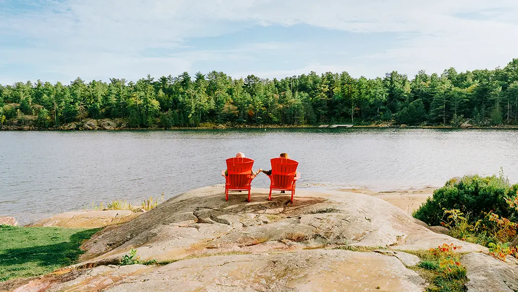 Two red chairs on a rocky shoreline overlooking a calm lake surrounded by forest.