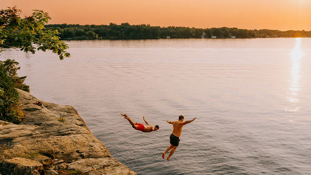 Deux personnes sautant dans un lac depuis une falaise rocheuse au coucher du soleil.