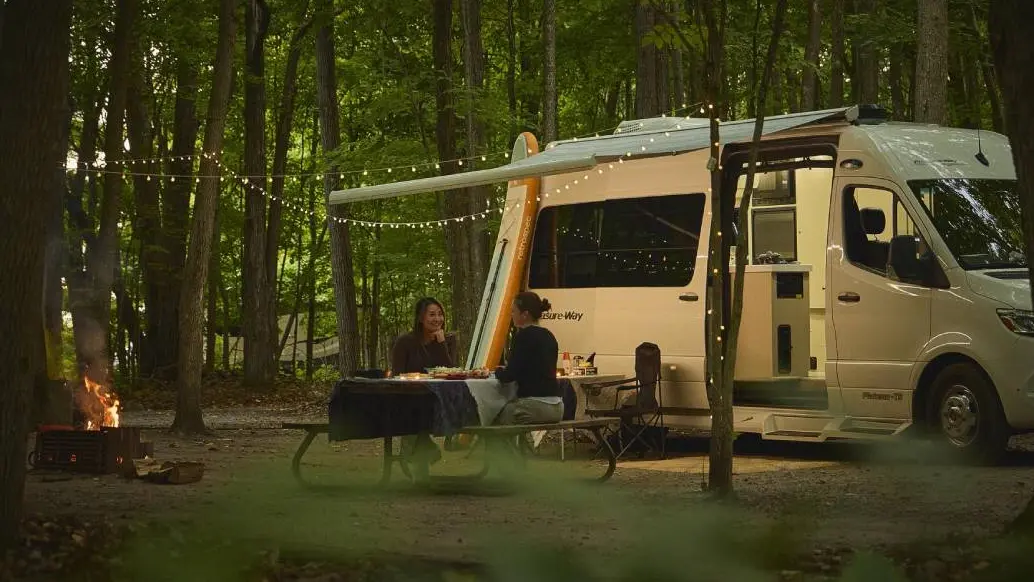 Dos mujeres disfrutan de una cena en una mesa de picnic junto a una caravana estacionada en un camping.
