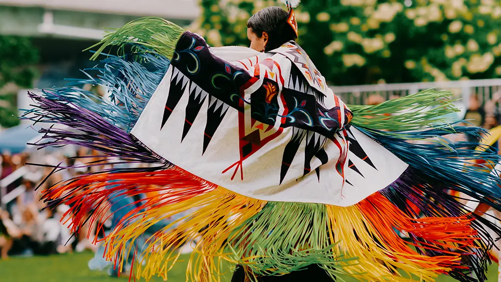 Indigenous dancer in vibrant traditional regalia with colourful fringe in motion during an outdoor powwow.