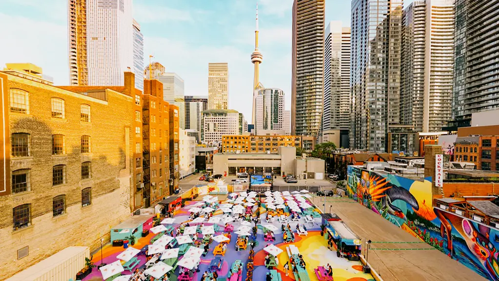Outdoor patio space with picnic tables, umbrellas and people gathered beneath a city skyline.