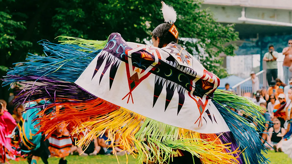 Bailarina indígena con un vibrante atuendo tradicional y coloridos flecos, en movimiento durante un powwow al aire libre.