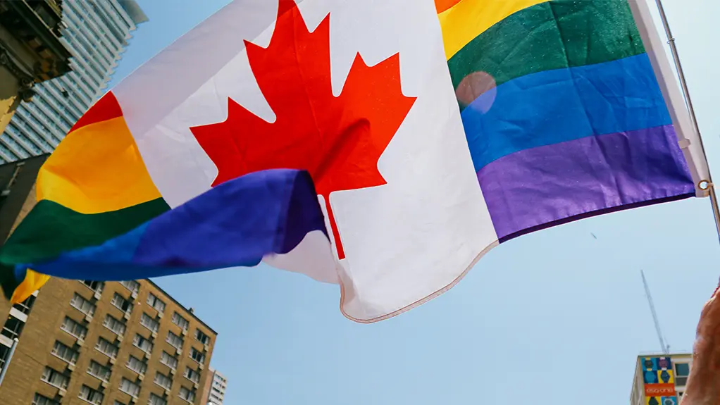 People waving rainbow and Canadian flags during the Toronto Pride parade on a city street with colourful festival crowds and sunlight flare.