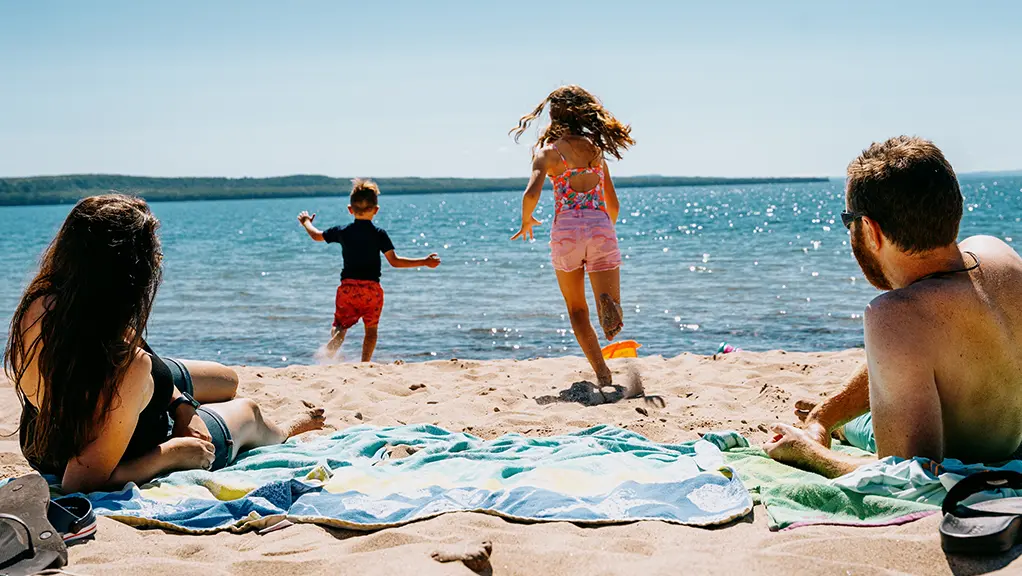 Two adults relax on towels at a sandy lakeside beach while two children run toward the water on a sunny day with clear blue sky.
