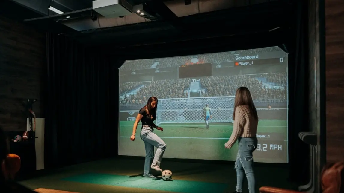 Un jugador tiene el pie sobre un balón de fútbol y está lanzando tiros a una portería virtual mientras otro jugador observa.