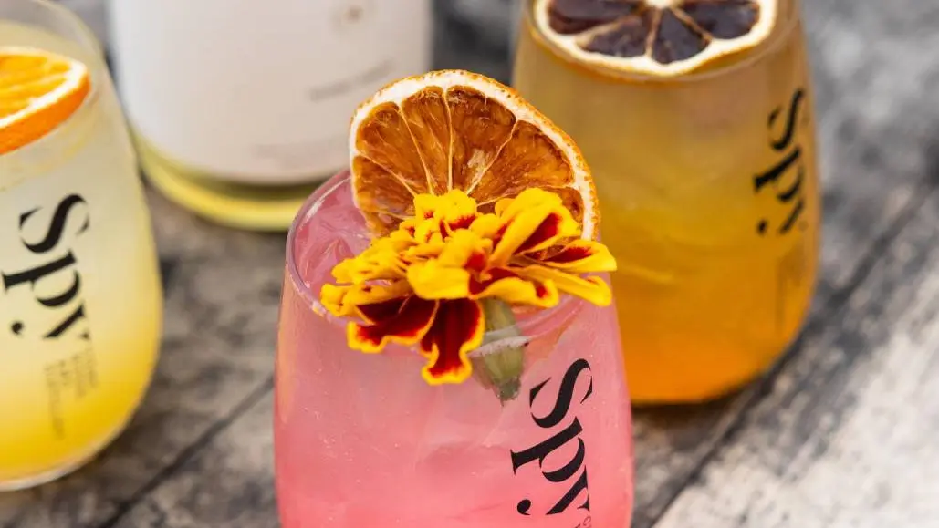 Trois verres de cidre coloré garnis de fruits et de fleurs posés sur une table en bois.