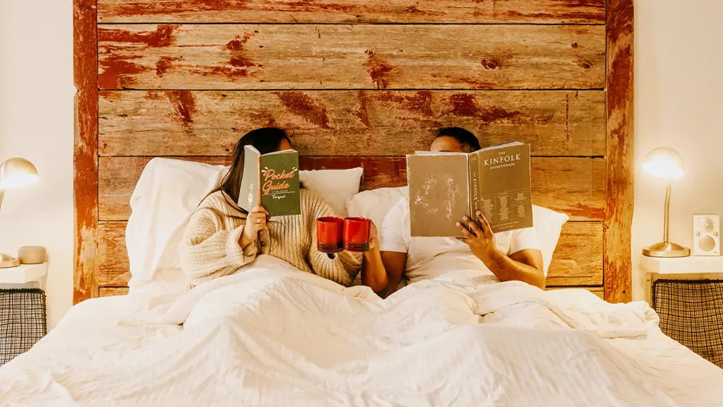 Un couple lit des livres au lit tout en tenant des tasses.