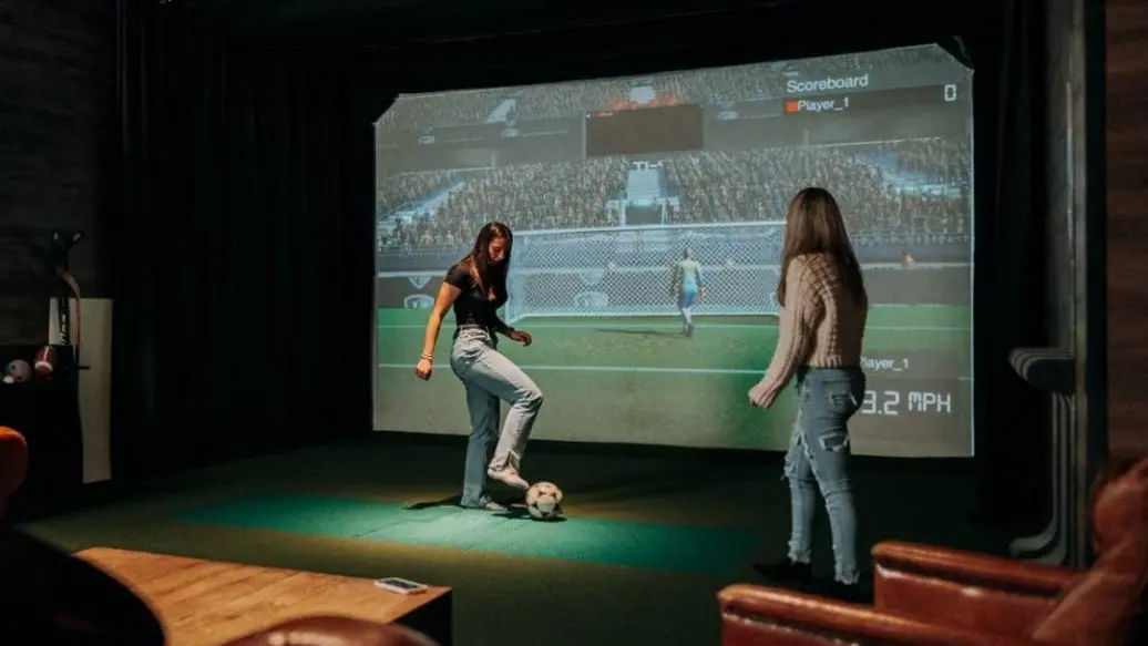 Un jugador tiene el pie sobre un balón de fútbol y está lanzando tiros a una portería virtual mientras otro jugador observa.