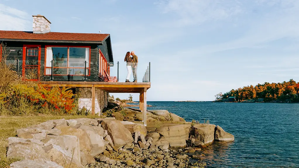 Ferienhaus am See mit Aussichtsplattform mit Blick auf ruhiges Wasser und ein bewaldetes Ufer.