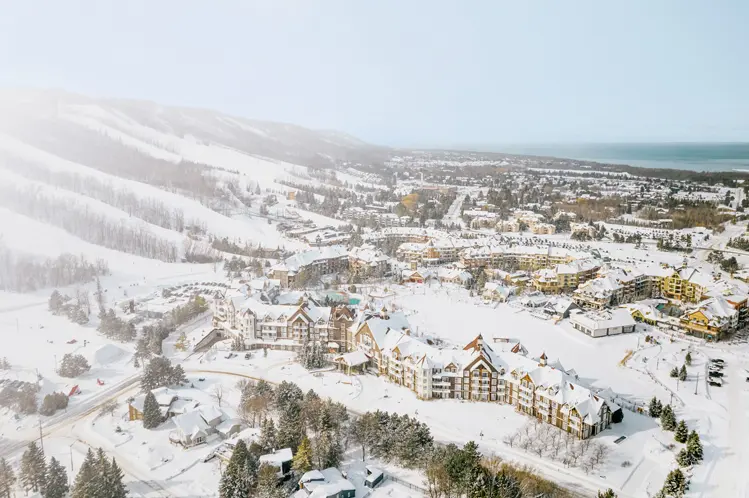 Vista aérea de un pueblo de esquí cubierto de nieve, con edificios de estilo chalet, pistas arboladas y la Bahía Georgiana visible en la distancia.